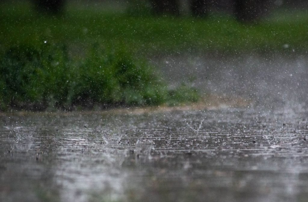 Chuva intensa atingindo o pavimento, com vegetação verde ao fundo.