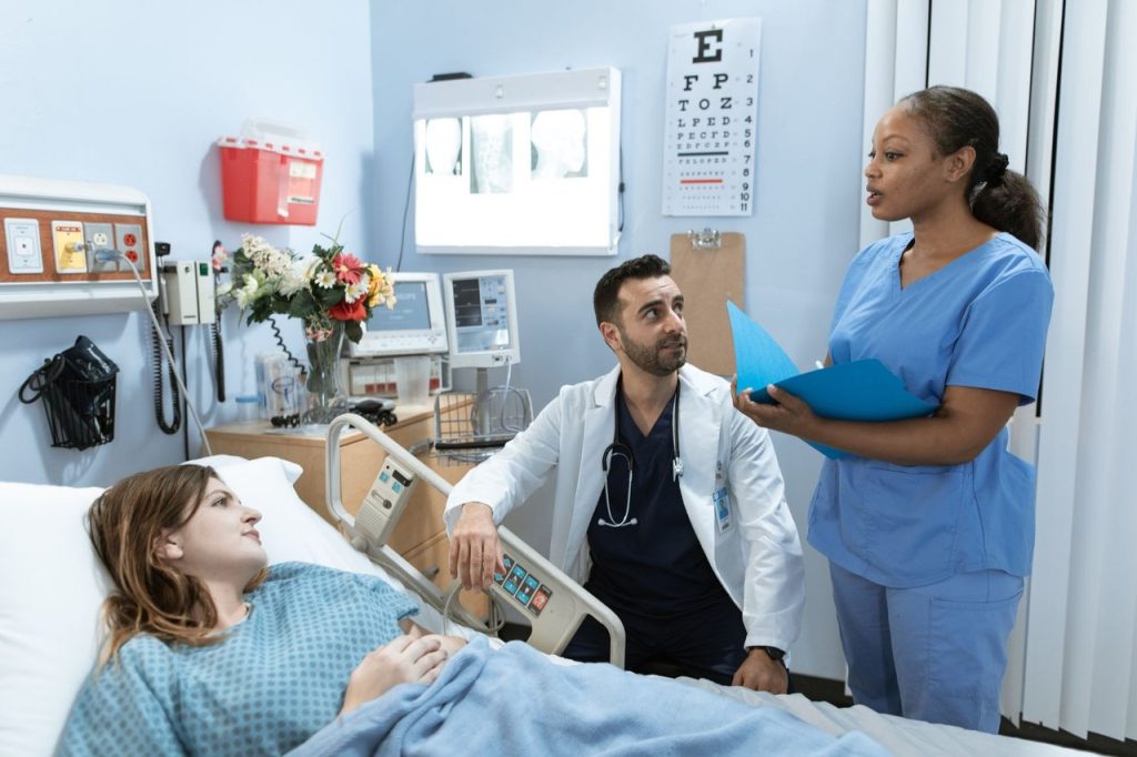 Médicos e enfermeira conversam sobre o tratamento de um paciente em um quarto hospitalar.